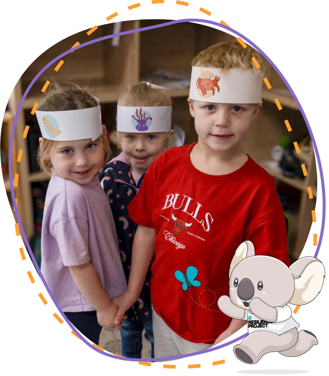 Three children wearing paper sea-creature headbands, holding hands in an early learning room